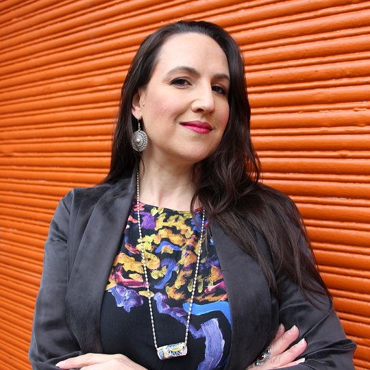 Photo of Dr Virginia Keft, she stands in front of an orange roller door, and has a smiling expression on her face, she has long dark straight hair and wears a black blazer with a couloiurful top underneath with yellow, pink, purple and blue . she has round silver dangling earrings, a large silver ring and a silver necklace with a cylindrical porcelain looking pendant